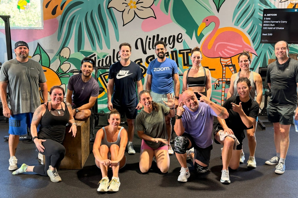 A group of fifteen people posing together in a gym in front of a colorful tropical-themed wall mural. The mural features palm leaves, a flamingo, and the text 'Flagler Village CrossFit'.